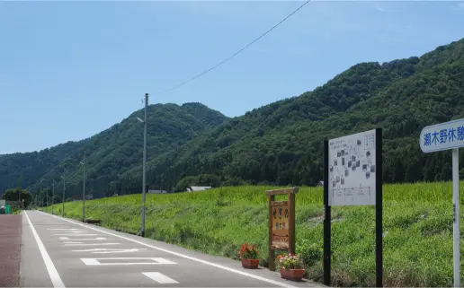A roadside rest area with signs and flowers along the Hakusan cycling path, surrounded by peaceful green hills.