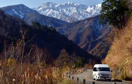 A scenic mountain road in the Hakusan area with a white van driving through colorful autumn foliage and snow-capped peaks in the background.