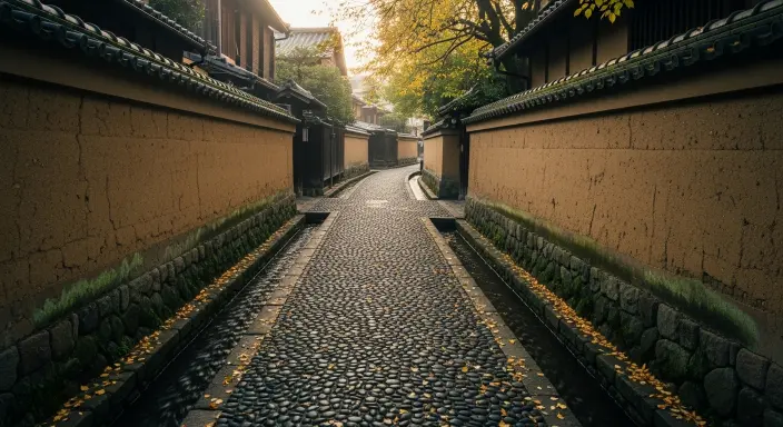 Stone-paved alley in Kanazawa’s Nagamachi Samurai District with earthen walls and small water canals, softly lit by morning light, evoking calmness and timeless beauty.