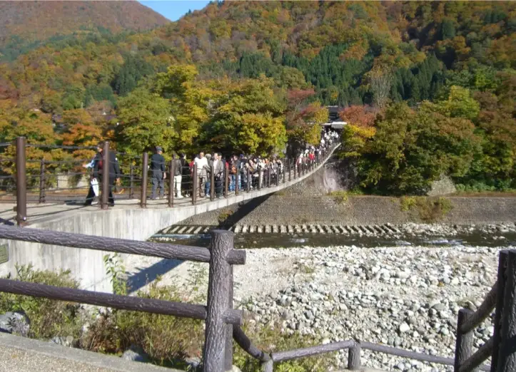 Crowds of visitors crossing Deai Bridge surrounded by vibrant autumn foliage and mountains in the UNESCO village of Shirakawago.