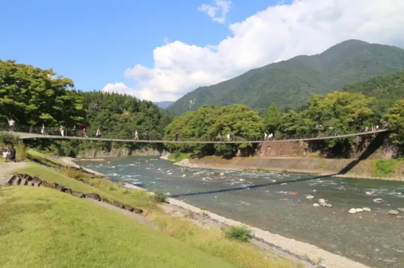 Panoramic shot of Deai Bridge stretching across the Shogawa River in Shirakawago, with lush green trees, clear blue sky, and mountains in the background.
