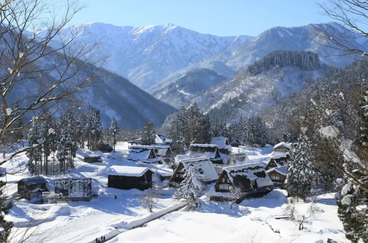 Panoramic winter view of Gokayama’s gassho-zukuri village blanketed in deep snow, with surrounding mountain ranges under a clear blue sky.