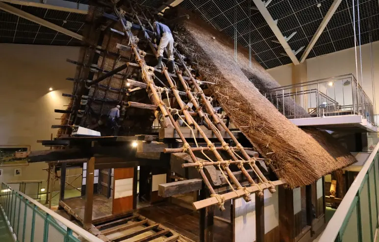 Indoor exhibit at Michi-no-Eki Shirakawa-go showing craftsmen repairing the steep thatched roof of a traditional gassho-zukuri house, with ladders and straw bundles in place.
