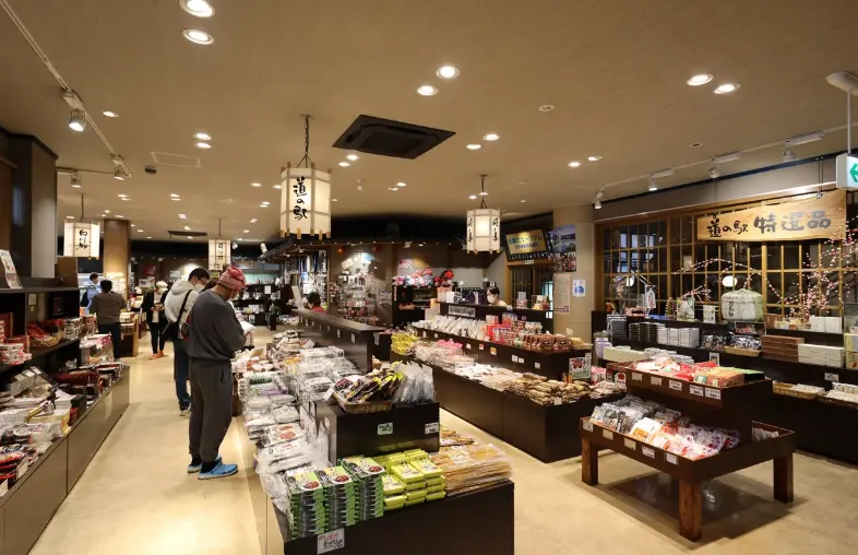 Bright, spacious interior of Michi-no-Eki Shirakawa-go’s souvenir shop displaying locally made snacks, crafts, and specialty products on wooden tables with visitors browsing.