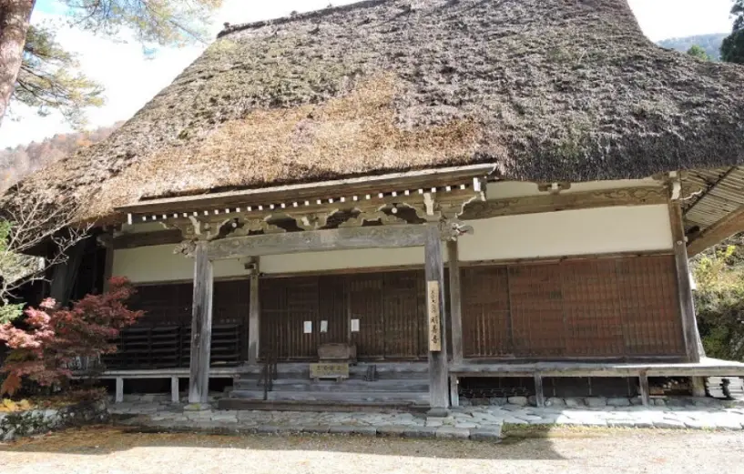 Front view of the main hall of Myosenji Temple featuring a massive thatched roof, wooden pillars, and autumn foliage around the historic building in Shirakawago.