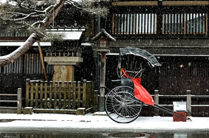 Snow-covered Sanmachi Suji in Takayama, with a traditional rickshaw standing in front of historic wooden buildings.