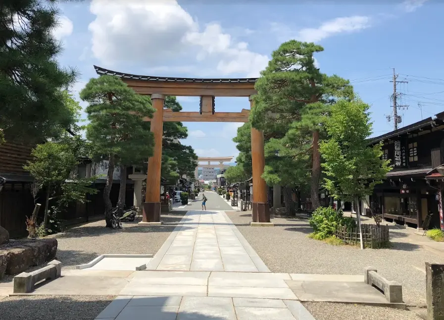 Sunny view of the main torii gate and approach leading to Sakurayama Hachimangu Shrine in Takayama, lined with pine trees and traditional wooden buildings.