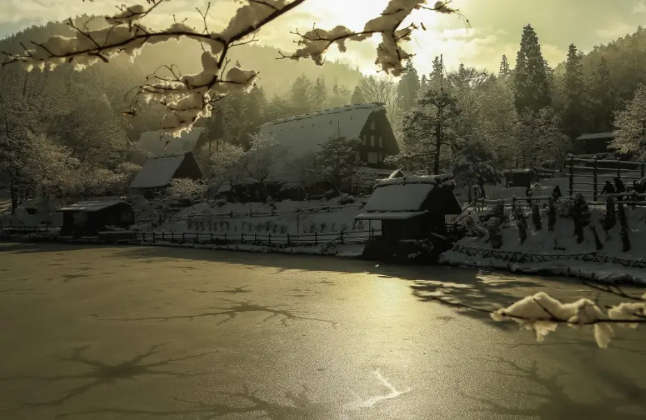 Snow-covered gassho-style houses at Hida Folk Village during winter, seen across a frozen pond in soft golden light.