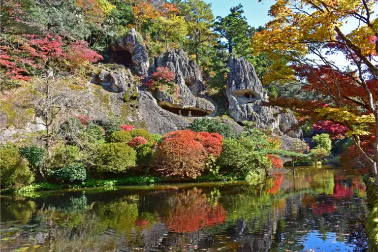 Scenic view of Natadera Temple's rock formations surrounded by colorful autumn leaves and reflected in a calm pond.
