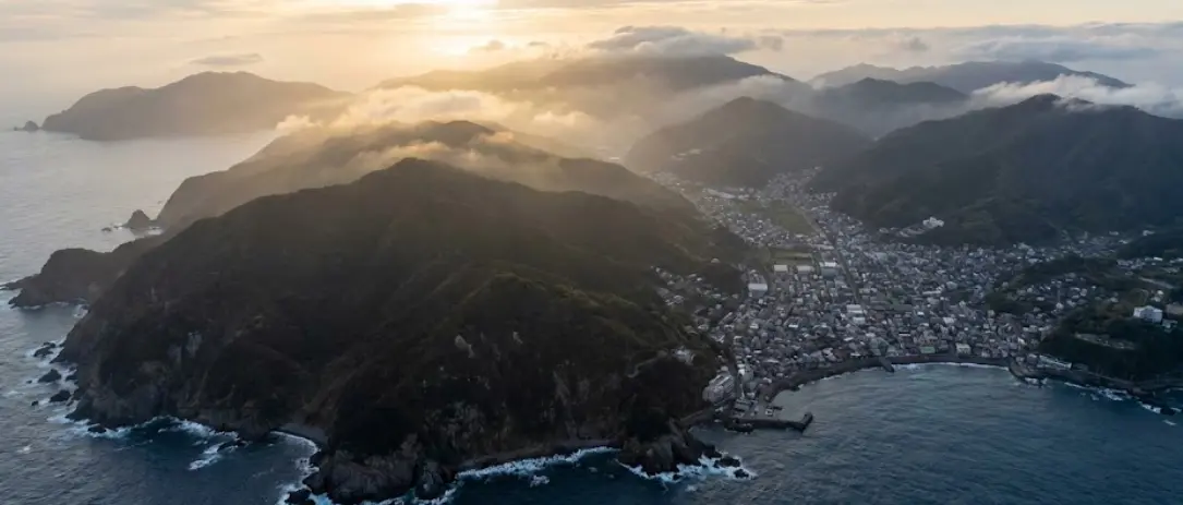 A coastal Japanese town nestled between steep forested mountains and the sea at sunset, illustrating limited land and dense settlement patterns.