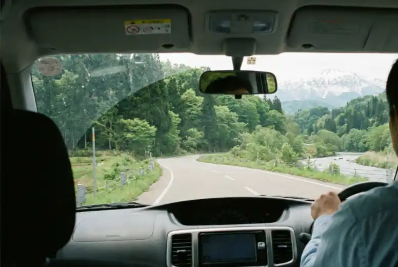 View from inside a car driving through the Hakusan area, with a winding road, river, forest, and snow-capped mountains in the distance.