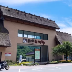 Front view of the Michi-no-Eki Shirakawa-go roadside station featuring a thatched-roof architectural style that echoes the gassho-zukuri houses of the region, set against green mountains and blue sky.