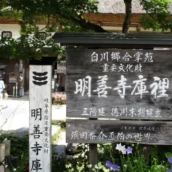 Wooden signboard marking the entrance to Myōzenji Kuri, a cultural heritage site in Shirakawago, surrounded by trees and traditional thatched buildings.